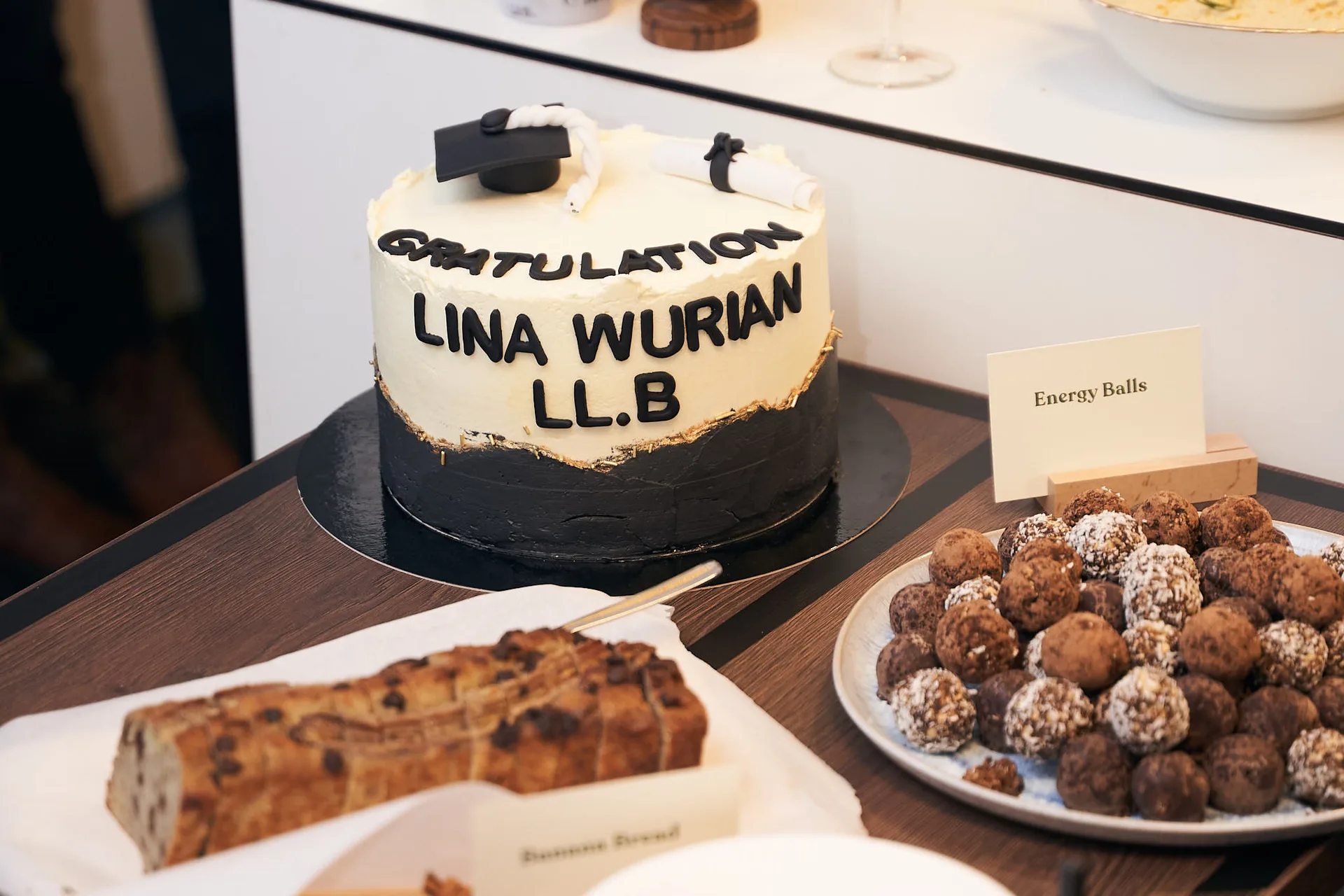 Eine zweifarbige Graduation-Torte mit weißer und schwarzer Creme, dekoriert mit essbarem Blattgold sowie einer Miniatur-Doktorhut-Figur und einem Schriftzug. Die Torte steht auf einem Buffet neben einer Platte mit „Energy Balls“ und einem Stück Bananenbrot.