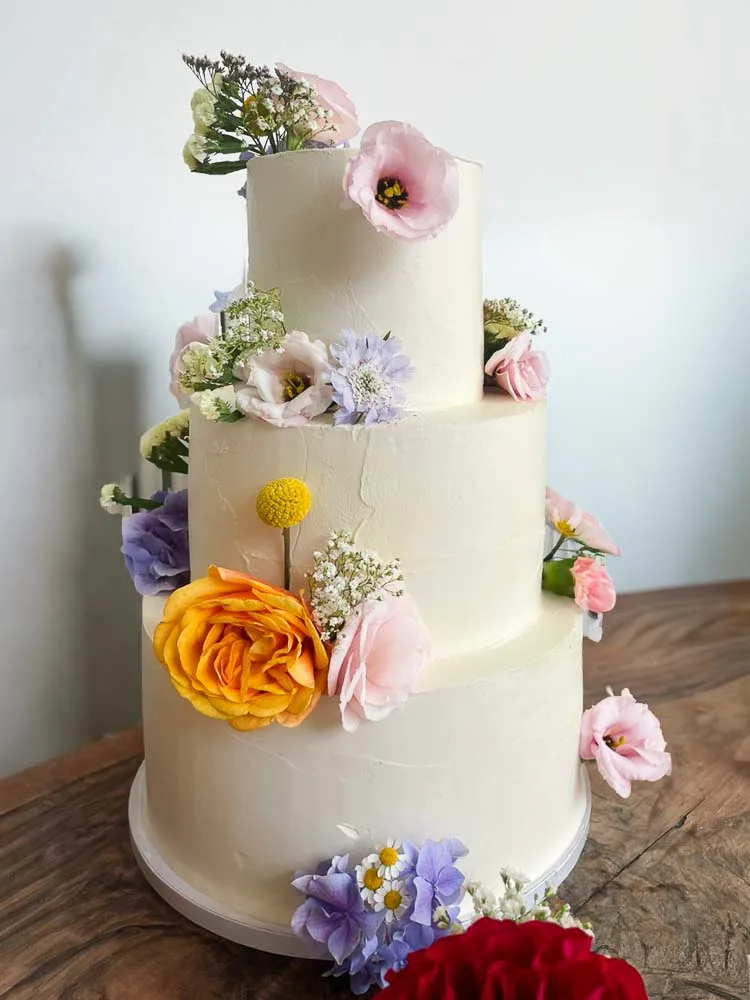 Dreistöckige, weiße Hochzeitstorte, üppig verziert mit verschiedenen bunten Blumen wie Rosen, Hortensien und Lisianthus in einer frühlingshaften Anordnung.