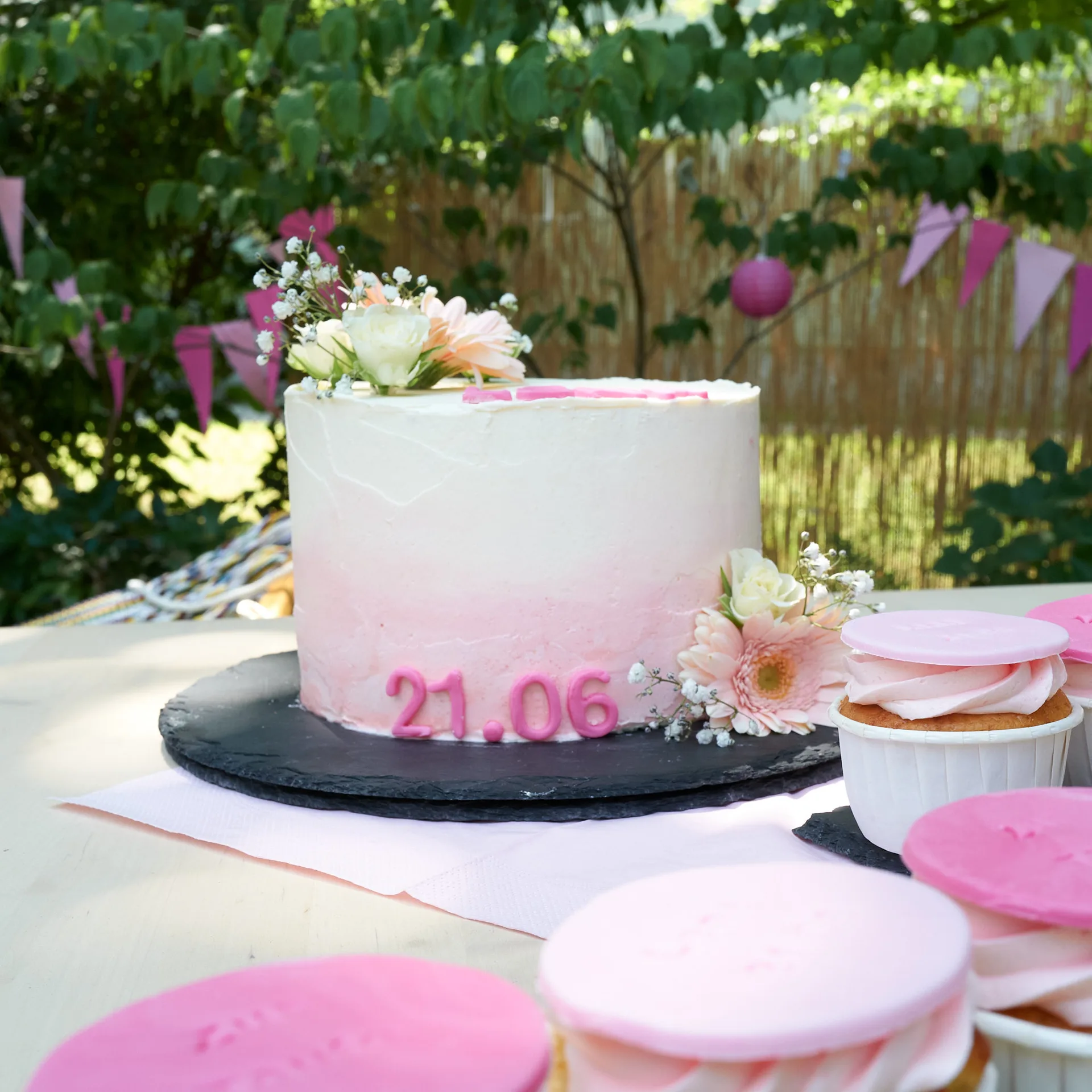 Eine festlich dekorierte Torte mit einem hellen Ombré-Farbverlauf von Weiß zu zartem Rosa, verziert mit frischen Blumen, weißer Creme und hellrosa Zuckerzahlen „21.06“ am unteren Rand. Im Vordergrund sind mehrere dazu passende Cupcakes mit rosa Zuckerhauben auf einem Tisch im Freien arrangiert.