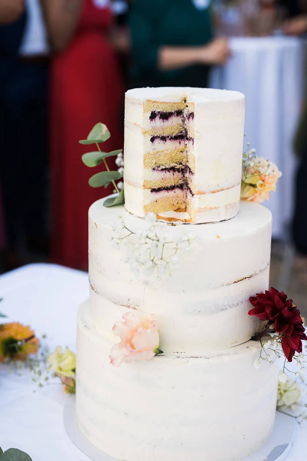 Dreistöckige, angeschnittene Hochzeitstorte im Semi-Naked-Cake-Stil, dekoriert mit Schleierkraut, Eukalyptus und frischen Blüten. Der Anschnitt zeigt mehrere Schichten Biskuit und dunkle Beerenfüllung.