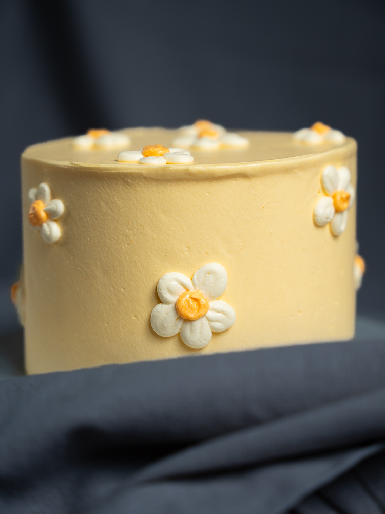 Torte 'Daisy' von oben und von der Seite fotografiert: Eine runde, pastellgelbe Torte mit handgefertigten weißen Margeriten-Verzierungen mit gelbem Kern auf einer Tortenplatte.