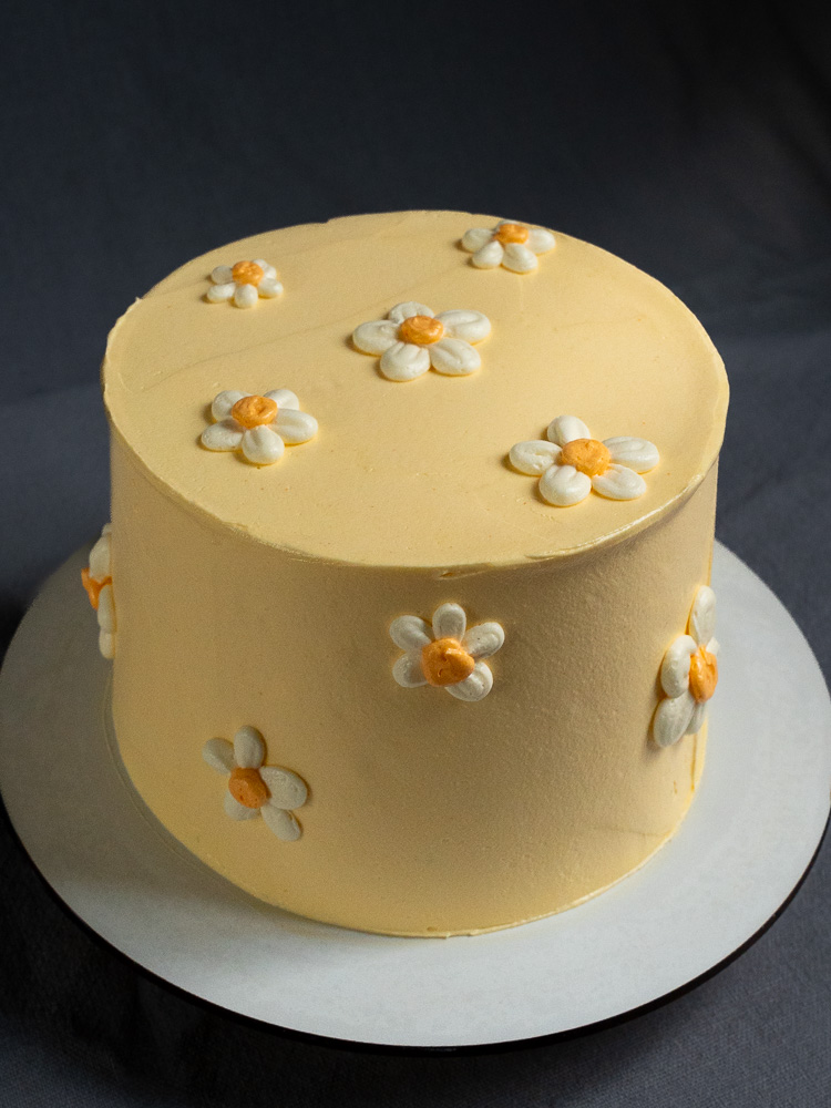 Torte 'Daisy' von oben und von der Seite fotografiert: Eine runde, pastellgelbe Torte mit handgefertigten weißen Margeriten-Verzierungen mit gelbem Kern auf einer Tortenplatte.